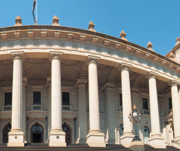 The steps and façade of Victoria's Parliament house on a sunny day. The Australian flag flies from the roof.