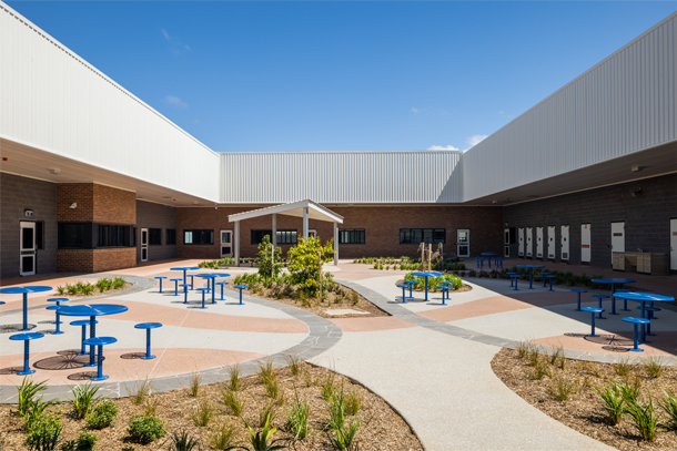 A photo of a courtyard in a Victorian prison. There are blue chairs and tables. The ground is paved. There are garden beds with native grasses.