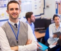 School principal facing camera in staffroom with other staff behind.