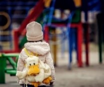 A young child with a fluffy yellow duck backpack watches an empty playground.