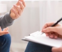 Close up of a patient speaking with their hands as a counsellor writes notes on a clipboard.