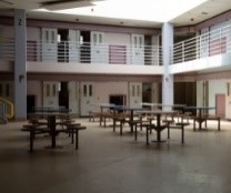 Tables and chairs in the common area of a prison. The common area is surrounded by numbered cells and there is light coming in through a skylight.