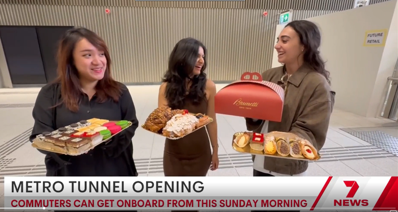 Screenshot of a 7 News television broadcast. The caption says ‘Metro tunnel opening. Commuters can get onboard from this Sunday morning’. Three people are smiling while holding trays of cakes and pastries.  