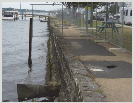 A hole in the path reveals a void behind the seawall where the bank has been eroded by water entering through gaps in the mortar. The wall's foundations have been undermined and the adjacent stormwater system (in the foreground) has failed.