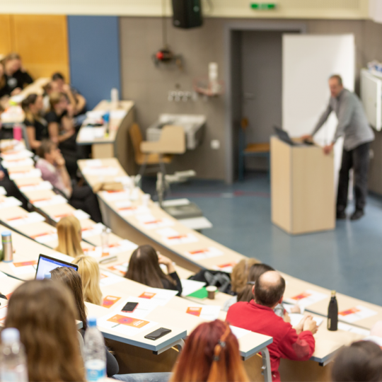 A small lecture theatre with three rows of students listening to a teacher speaking behind a podium.