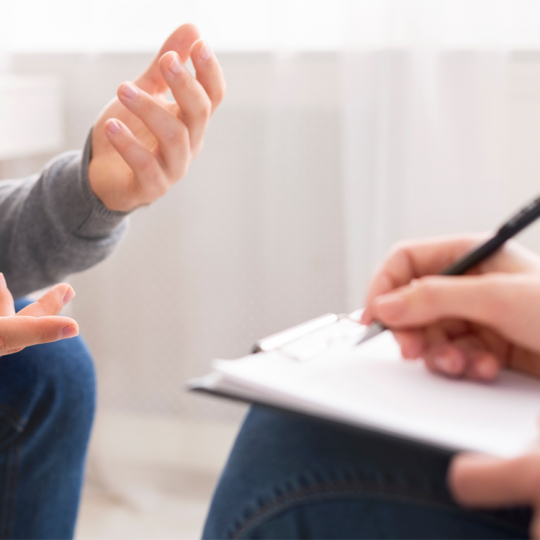 Close up of a patient speaking with their hands as a counsellor writes notes on a clipboard.