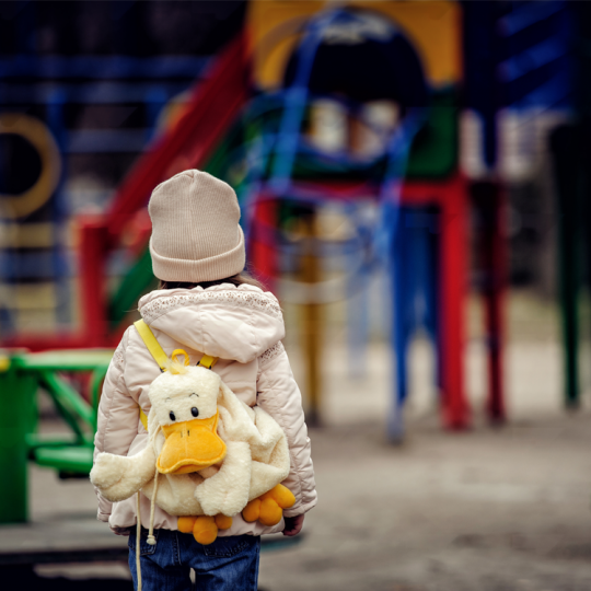 A young child with a fluffy yellow duck backpack watches an empty playground.