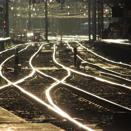 Multiple rows of metal railway tracks glowing in the sun. There are signals and other train infrastructure in the background.