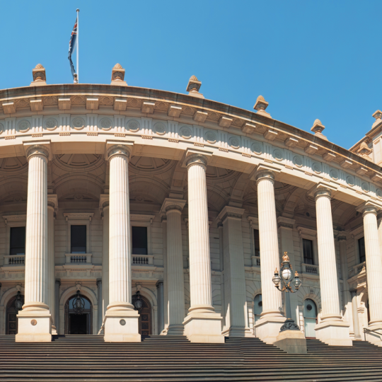 The steps and façade of Victoria's Parliament house on a sunny day. The Australian flag flies from the roof.