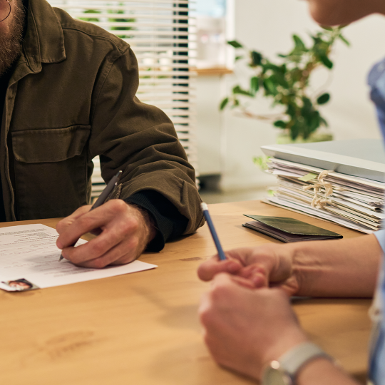 Two people sitting at a table. There are documents on the table and one person is filling a form.