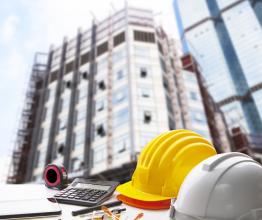 Image shows safety helmet and writing instrument on engineering working table against exterior construction building. Photo courtesy of stockphoto mania/Shutterstock.com