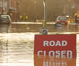 Image shows a 'Road Closed' sign partially covered in flood water lit by the evening sun. Photo courtesy of AC Rider/Shutterstock.com