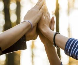 Image shows four female hands joined together high up in the air outside in a woodland. Photo courtesy of Gajus/Shutterstock.com