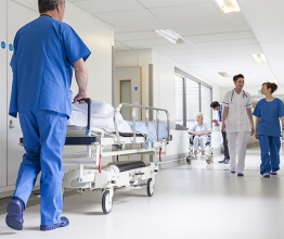 Male nurse pushing a hospital bed down a corridor in a hospital. Two other nurses are walking towards him.