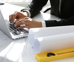 Cropped view of young Caucasian engineer wearing formal suit working on construction project using laptop computer, sitting at office desk, surrounded with engineering tools and rolls of blueprints. Photo courtesy of WAYHOME studio/Shutterstock.com