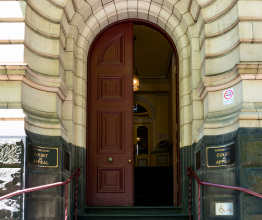 Doorway and entrance to the Supreme Court of Victoria. The bottom half of the doorway and the stairs leading up to it are made from sandstone. The upper doorway arch and walls are made of sandstone and the door is maroon and half open.