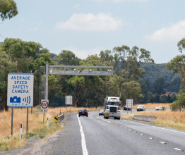 Cars and trucks driving past an average speed safety camera on a rural highway.