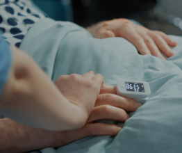 Close up of a nurse's hand holding a patient's hand. The patient is in a hospital bed and wearing a digital monitoring device on their finger.