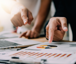 Close up of two people's hands pointing to a clipboard on a desk. The clipboard has a piece of paper with colourful graphs on it. There is a laptop in the background.