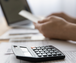 Black calculator sitting on top of accounting files on a desk. A person's hands holds an out-of -focus piece of paper in front of a laptop in the background.