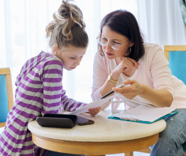 A woman and a young girl sitting at a small table and reading a document together.