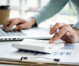 Close up of a woman's hands using a calculator and laptop. The calculator is sitting on a clipboard with accounting documents.