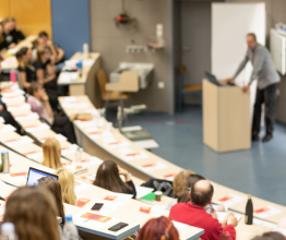 A small lecture theatre with three rows of students listening to a teacher speaking behind a podium.