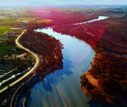 Aerial photo of a river in a regional area. The river is surrounded by bushland. There is a road winding beside the river and farmland in the distance.