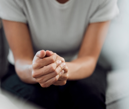 A close-up photo of a woman sitting on a couch with her hands clasped together on her knees.