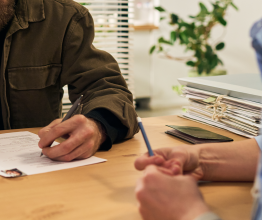 Two people sitting at a table. There are documents on the table and one person is filling a form.